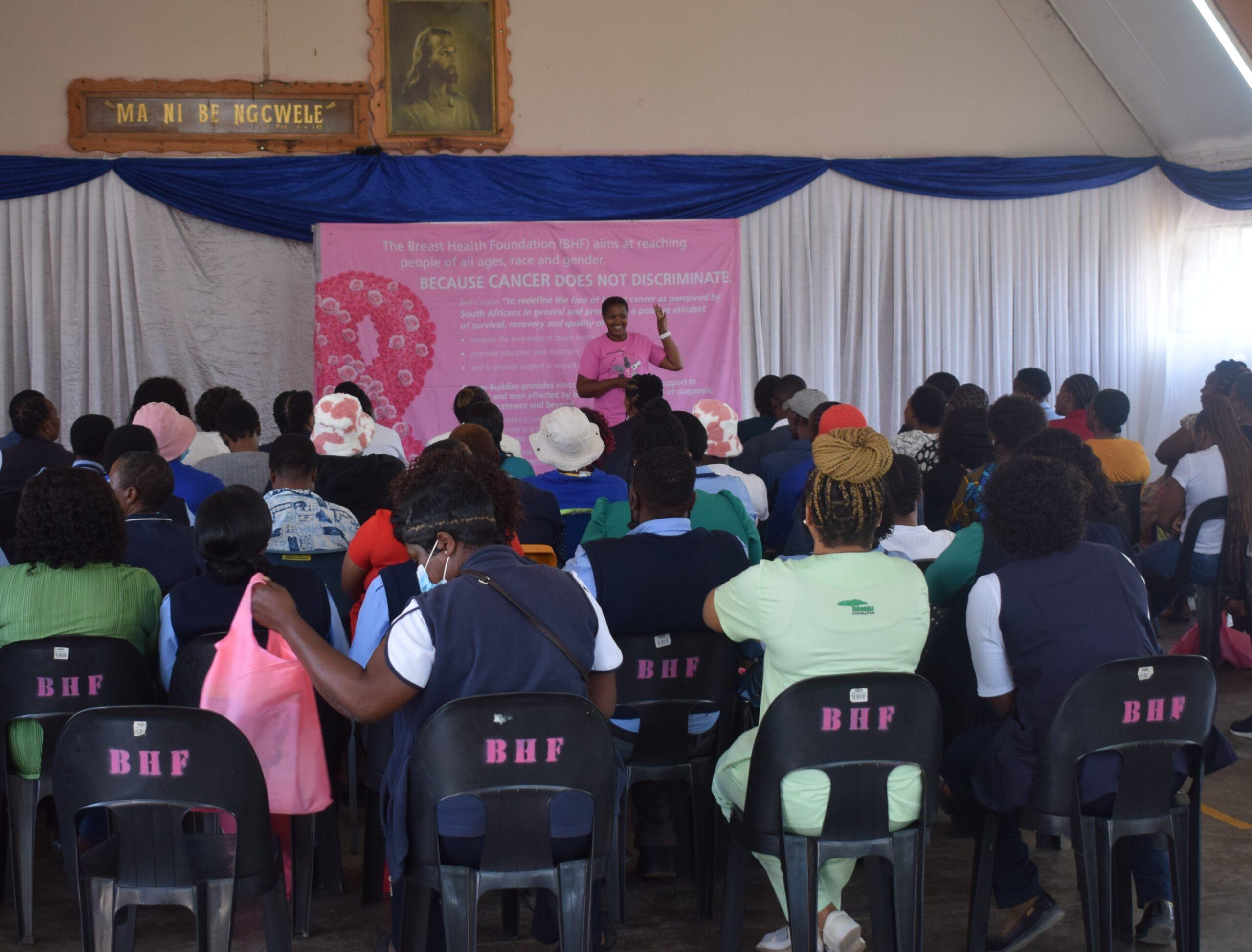 A Breast Health Foundation survivor Navigator in pink presenting a breast health awareness talk to community members seated in a clinic.